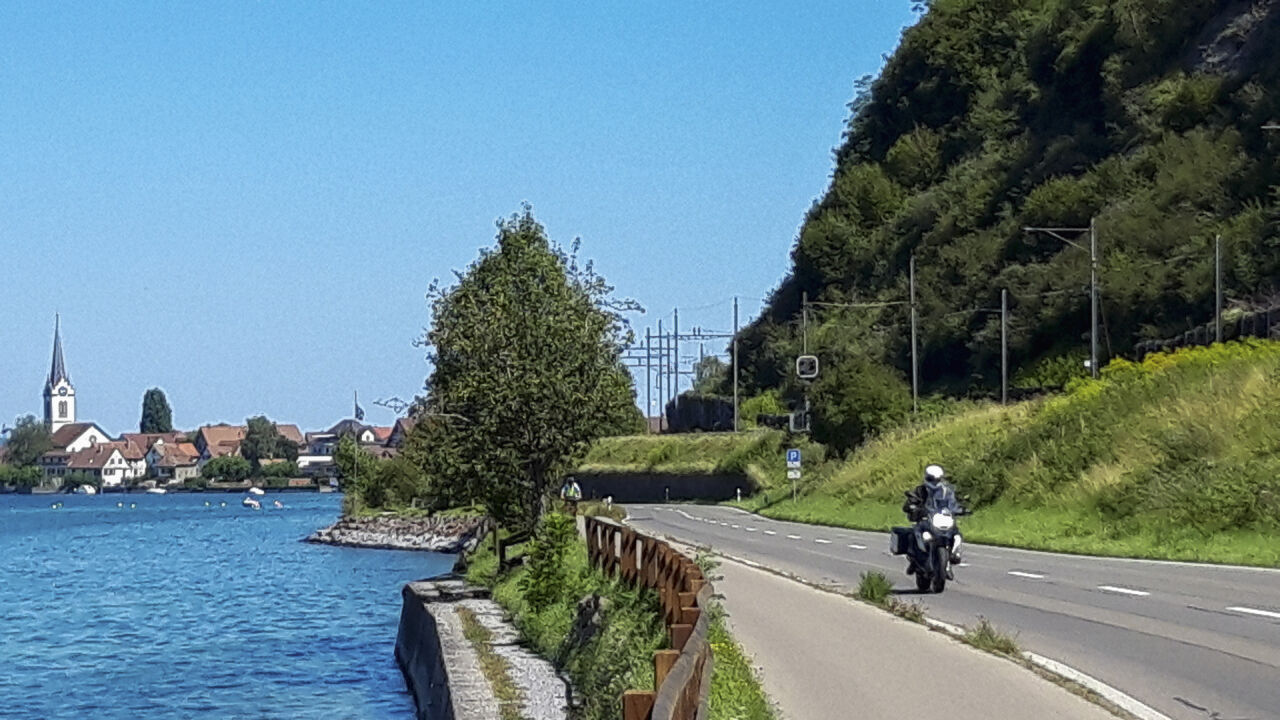 Motorrad auf einer Uferstraße, links der See, im Hintergrund ein Dorf mit Kirchturm Motorrad auf einer Uferstraße, links der See, im Hintergrund ein Dorf mit Kirchturm