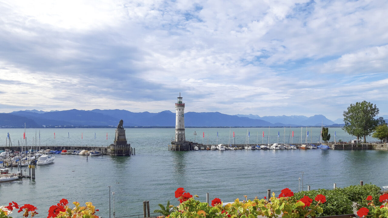 Hafeneinfahrt mit Leuchtturm und Löwenstatue auf der Mole, dahinter Berge über dem See Hafeneinfahrt mit Leuchtturm und Löwenstatue auf der Mole, dahinter Berge über dem See